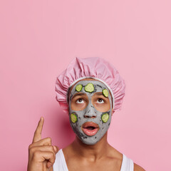 Vertical shot of stunned African man concentrated above applies nourishing clay mask and cucmber for skin moisturising and reducing fine lines shows blank space for your advertising content.