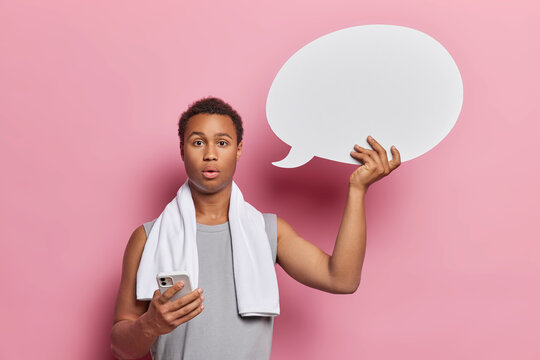 Shocked dark skinned man finds pleasure in his home workout routine poses with towel around neck holds smartphone and blank speech bubble for your advertising content isolated over pink background