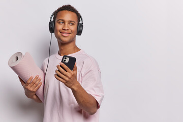 Studio shot of sporty young man holds mobile phone enjoys listening music via headphones holds rolled karemat prepares for yoga or fitness training looks gladfully aside isolated over white background