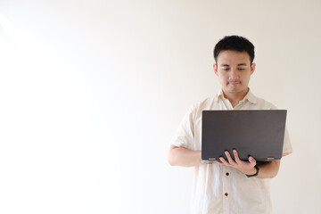 A young Asian man wearing a beige shirt and a smartwatch on his left wrist is using a black laptop withstanding and serious facial expression. Isolated white background. Suitable for advertisement.