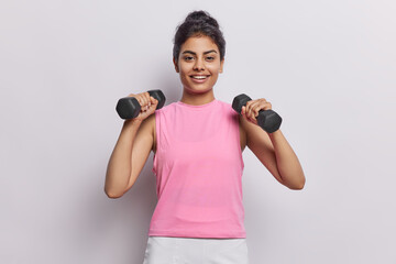 Horizontal shot of sporty motivated woman raises her arms while performing fitness exercises with dumbbells showcases strength and determination dressed in sportswear isolated over white background