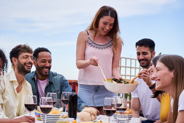 A caucasian young woman gathered with her friends, serves salad to the guests. Group of cheerful young adults hanging out together in a house terrace, eating food and drinking wine.