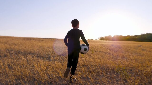 A boy with a soccer ball in his hands runs through a meadow at sunset. The concept of a kids dream, sports, forward. Silhouette of a child at sunset with a ball