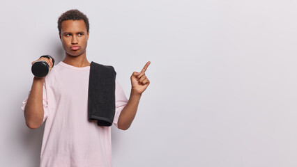 Waist up shot of serious African man engages in intense physical exercises at gym holds dumbbell points finger at empty space for your promotional content wears t shirt isolated over white background