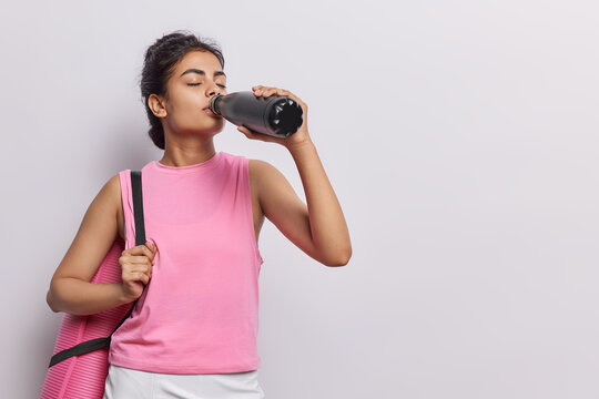 Horizontal shot of active woman rests after yoga session carries rolled karemat and drinks water feels thirsty dressed in pink t shirt stands against white background empty space for your advert