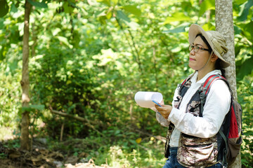 Asian woman botanist is at forest, hold paper notepad to survey  botancal plants in forest. Concept,  field research outdoor.                                  Nature surveying. Ecology and environment - obrazy, fototapety, plakaty