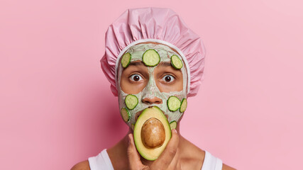 Shocked African man applies beauty facial mask with cucumber slices for skin moisturising covers mouth with half of avocado wears bath hat isolated on pink background. Wellness and cosmetology