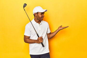 African American man golfer, yellow studio backdrop, showing a copy space on a palm and holding another hand on waist.