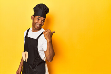 African American cook with utensils, yellow studio, points with thumb finger away, laughing and carefree.