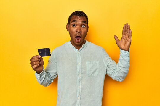 African American man with credit card, yellow studio, surprised and shocked.