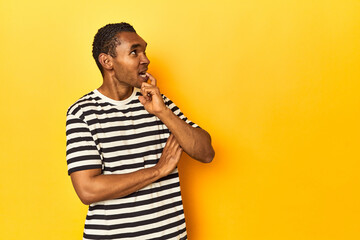 African American man in striped t-shirt, yellow studio, looking sideways with doubtful and skeptical expression.