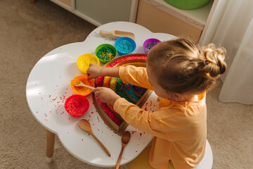 A little girl playing colored rice and make rainbow. Child filled the rainbow with bright rice. Montessori material. Sensory play and learning colors activity for kids. - obrazy, fototapety, plakaty