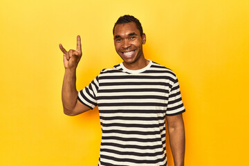 Fototapeta premium African American man in striped t-shirt, yellow studio, showing a horns gesture as a revolution concept.