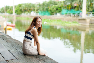 Fototapeta na wymiar Asian cute teenage girl with long hair Sit and relax in your free time on the pier. By the river on weekends with a beaming face. A rural canal with clear water