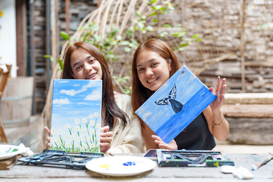 Portrait. Two Asian female apprentice painters sitting and holding paintings in hands. Art teacher's watercolor painting with brush, a whale, the sea and field of flowers in school's art classroom. - Powered by Adobe