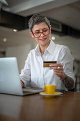 One senior woman with eye glasses holding credit card and shopping online on laptop, modern consumer concept