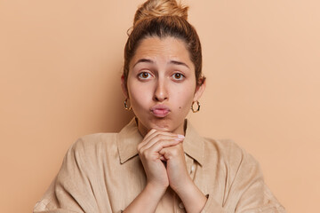 Portrait of sad lovely European woman feels disappointed keeps lips folded hands under chin wears shirt concentrated at camera isolated over brown background. Human facial expressions concept