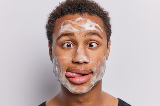 Close up shot of funny guy adiates joy as he indulges in pampering session gently applying refreshing bubble cleansing foam sticks out tongue and crossed eyes isolated over white background.