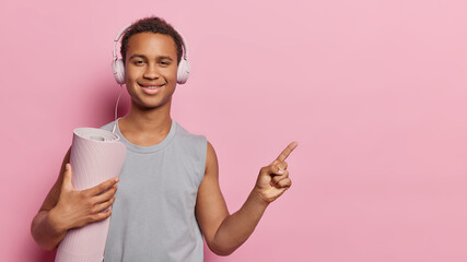 Horizontal shot of handsome sporty man dressed in grey t shirt poses with rolled karemat listens favorite music via headphones points index finger aside on blank space isolated over pink background