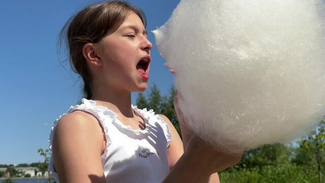 a happy brunette girl appetizingly eats sweet cotton candy against a blue sky background, a child eats cotton candy on the street and licks his fingers