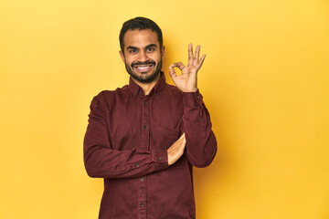 Casual young Latino man against a vibrant yellow studio background, winks an eye and holds an okay gesture with hand.