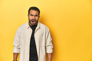 Casual young Latino man against a vibrant yellow studio background, shouting very angry, rage concept, frustrated.