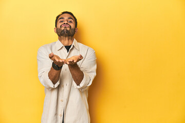 Casual young Latino man against a vibrant yellow studio background, folding lips and holding palms to send air kiss.