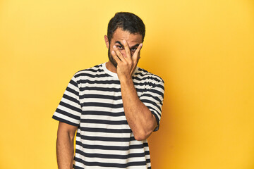 Casual young Latino man against a vibrant yellow studio background, blink at the camera through fingers, embarrassed covering face.