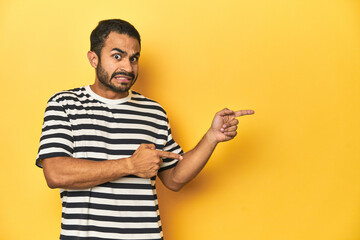 Casual young Latino man against a vibrant yellow studio background, shocked pointing with index fingers to a copy space.