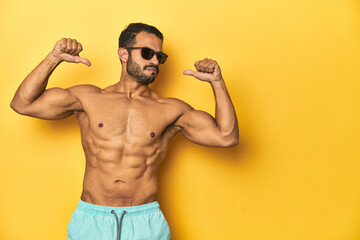 Fit young Latino man in swimwear and sunglasses, yellow studio background, feels proud and self...