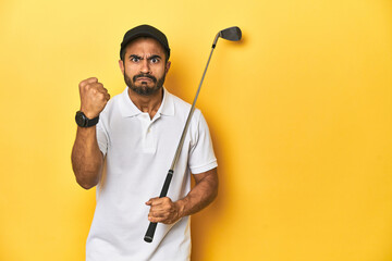 Young Latino golfer with club and cap on a yellow studio background, showing fist to camera,...