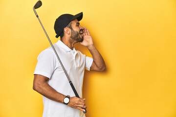 Young Latino golfer with club and cap on a yellow studio background, shouting and holding palm near opened mouth.