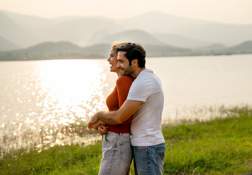 Caucasian man hug woman from the back and stay together near lake with sunset and romance in area of public park.
