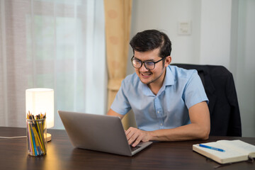 Asian young men in glasses is using laptop working at desk happily at home.