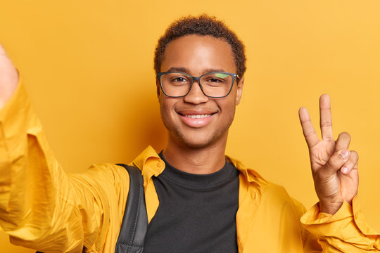Handsome young man dressed in shirt showcases his beaming smile as he takes selfie gracefully gesturing peace spreading positivity through social media carries backpack isolated over yellow background