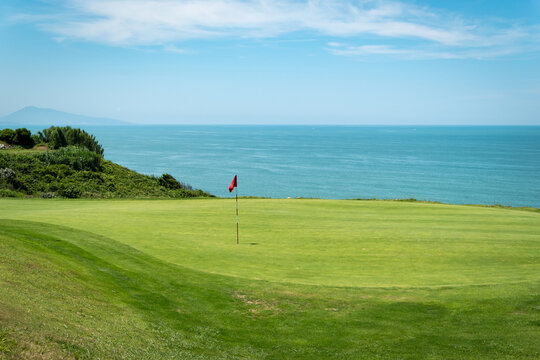 Campo de golfe com uma bandeira vermelha a meio e o mar ao fundo num dia ensolarado