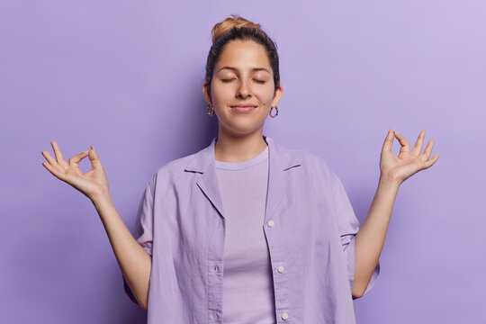Calm relaxed woman finds tranquility through mindful meditation her hands form serene mudra gesture wears shirt keeps eyes closed isolated over purple background practices yoga after hard day