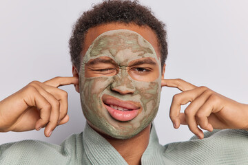 Close up portrait of African man applies clay mask and using beauty products or cosmetics highlighting essence of skin care and peeling in professional setting plugs ears poses indoor alone.