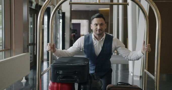 Dolly shot of helpful male porter wearing stylish uniform moving luggage cart along hotel lobby. Guest service and occupation concept.