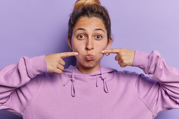 Photo of funny young European woman blows cheeks pouts lips points index fingers at face holds...