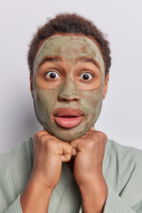 Shocked young man has face covered with clay mask wants to have healthier skin seeking solutions for skincare problems keeps hands under chin wears bath robe isolated over white studio background