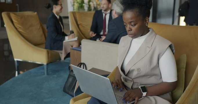 Successful African American businesswoman is working with laptop in hotel lobby concentrated on online activity busy with networking.