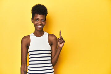 Vibrant African woman, white tank-top, yellow studio, showing number one with finger.