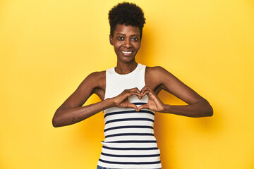 Vibrant African woman, white tank-top, yellow studio, smiling and showing a heart shape with hands.