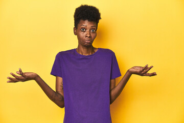 African woman in violet tee, yellow studio scene, doubting and shrugging shoulders in questioning...