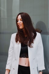 
beautiful young woman in a white jacket and black trousers with an open belly and a tattoo in the city against the backdrop of glass buildings