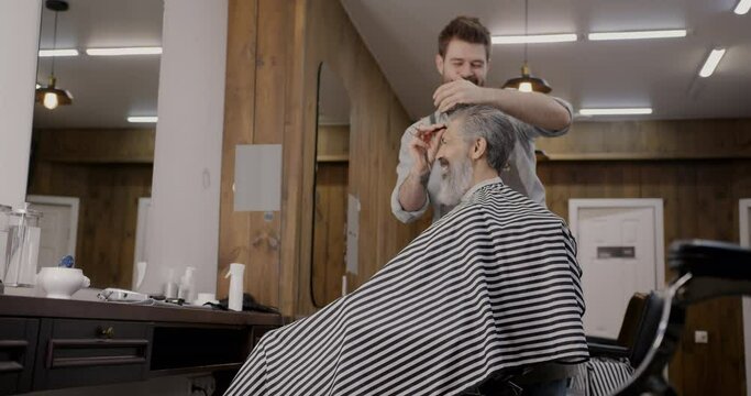 Barber working with client combing gray hair talking and laughing indoors in barbershop. Professional occupation and beauty salon concept.