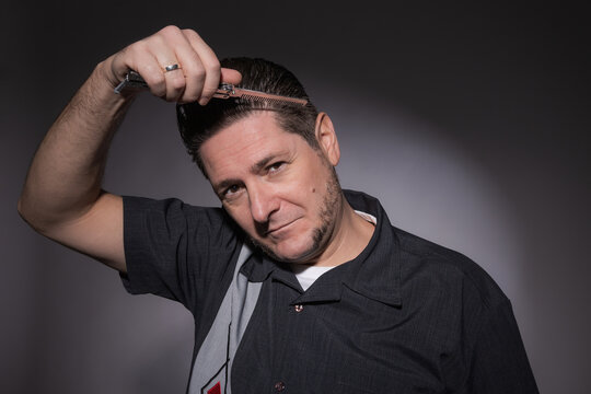 Horizontal studio portrait of a 40 year old man in rockabilly style combing his toupee with a steel folding butterfly comb