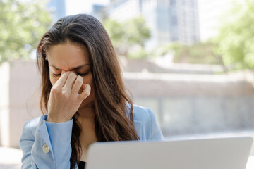 Tired middle aged latin businesswoman using laptop computer rubbing eyes, working online at street. Stressed woman having headache while searching information. Frustrated freelancer missed deadline 