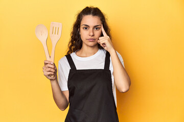 Woman with apron, wooden cooking utensils, yellow, pointing temple with finger, thinking, focused...